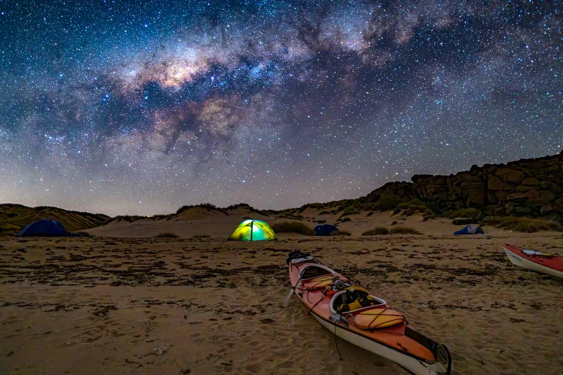 An illuminated tent under the vibrant Milky Way, with a sea kayak in the foreground, in the Dampier Archipelago