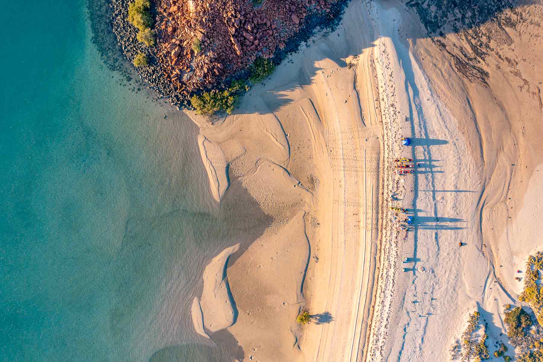 An aerial image of sea kayaks and people on a beach fringed by red rocks and turquoise water 